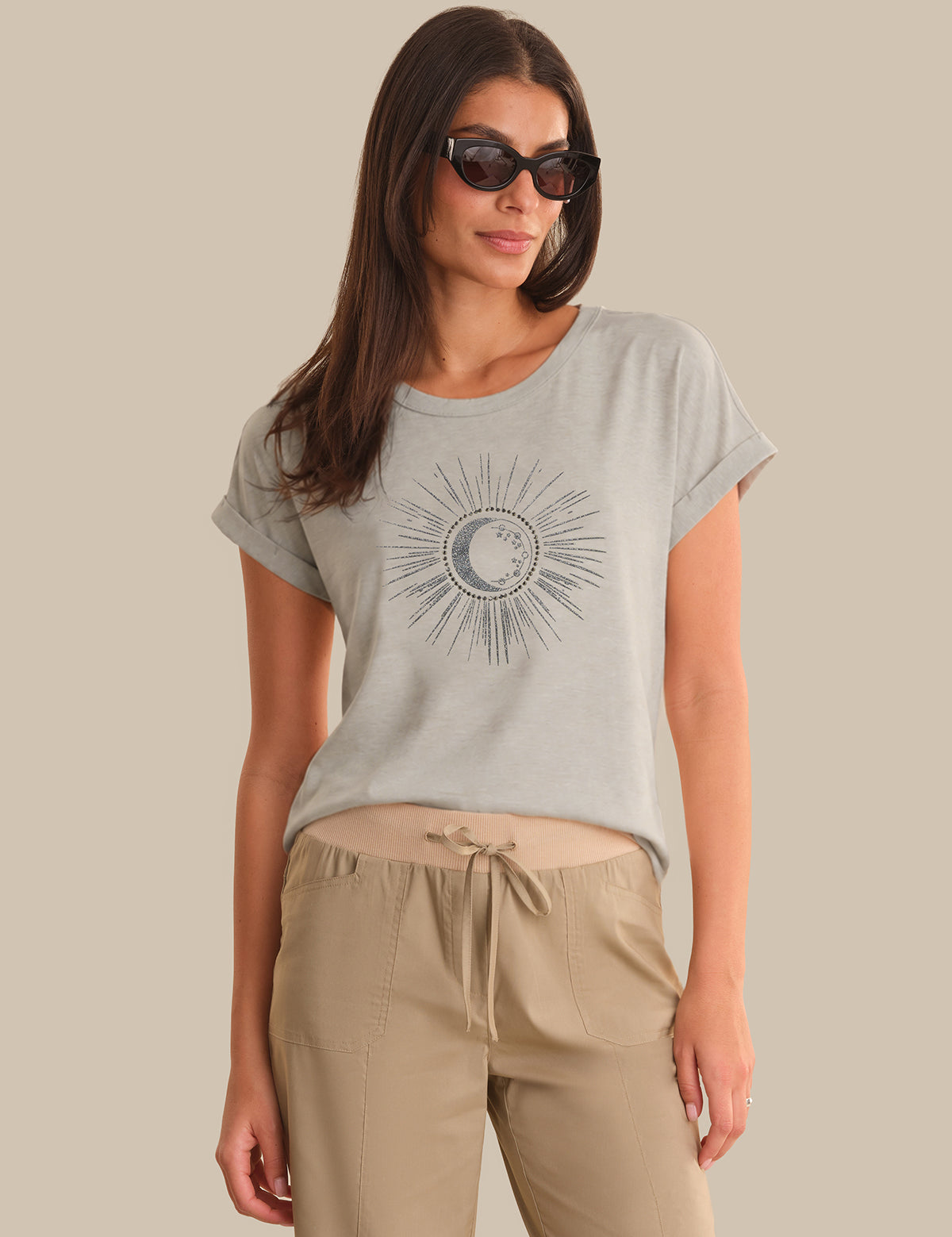Woman wearing a light gray t-shirt with a sun and moon design and beige pants against a beige background.