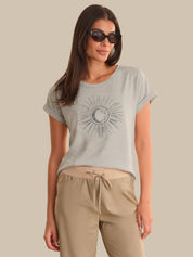 Woman wearing a light gray t-shirt with a sun and moon design and beige pants against a beige background.