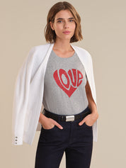 Woman wearing a gray t-shirt with a red heart and 'LOVE' text, paired with a white blazer and dark pants on a beige background.