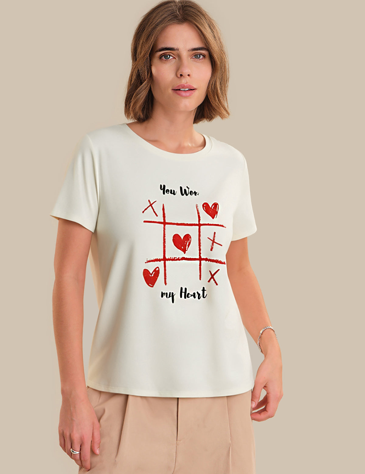 Woman wearing a white t-shirt with a red heart and cross-out design, standing against a beige background.