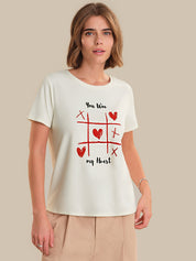 Woman wearing a white t-shirt with a red heart and cross-out design, standing against a beige background.