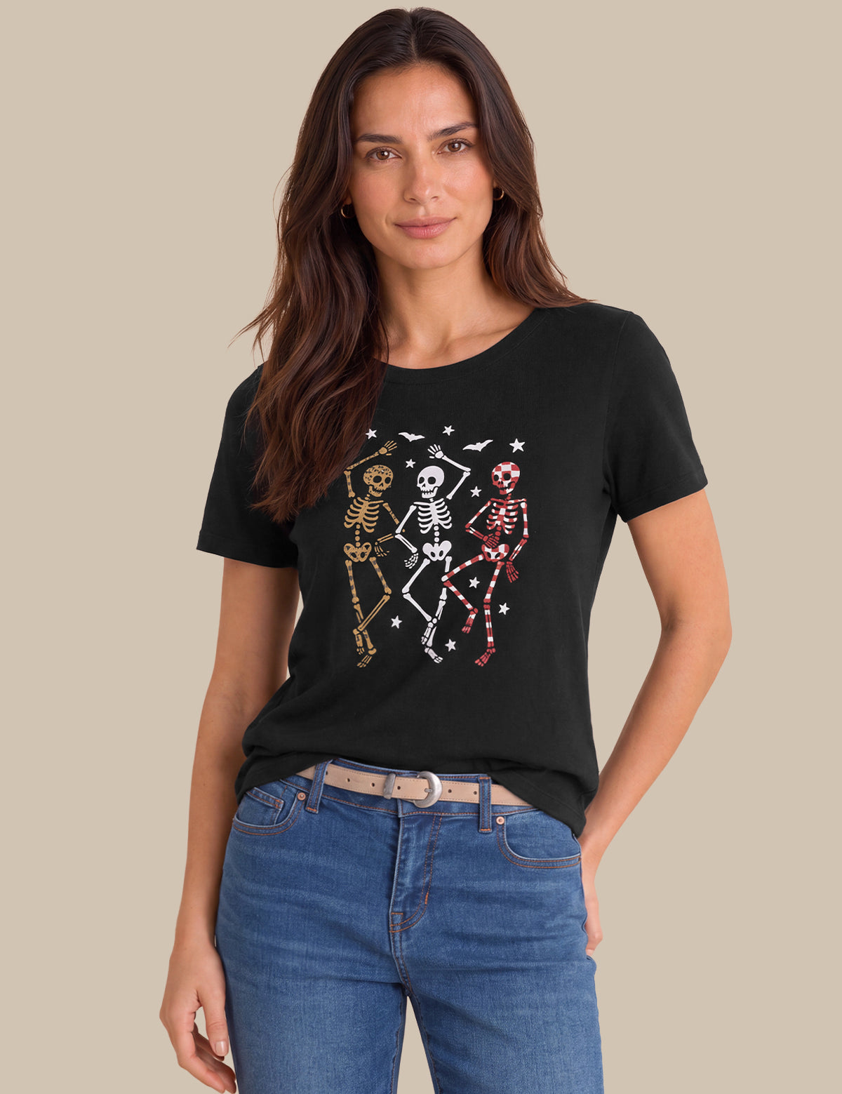 A woman modeling a black short-sleeve t-shirt with a graphic of three dancing skeletons in gold, white, and pink, paired with blue jeans against a solid beige background.