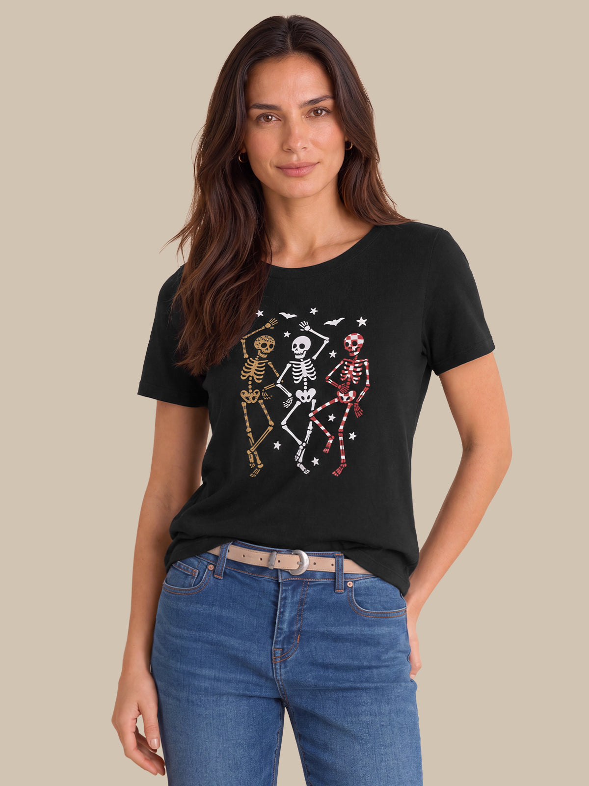 A woman modeling a black short-sleeve t-shirt with a graphic of three dancing skeletons in gold, white, and pink, paired with blue jeans against a solid beige background.