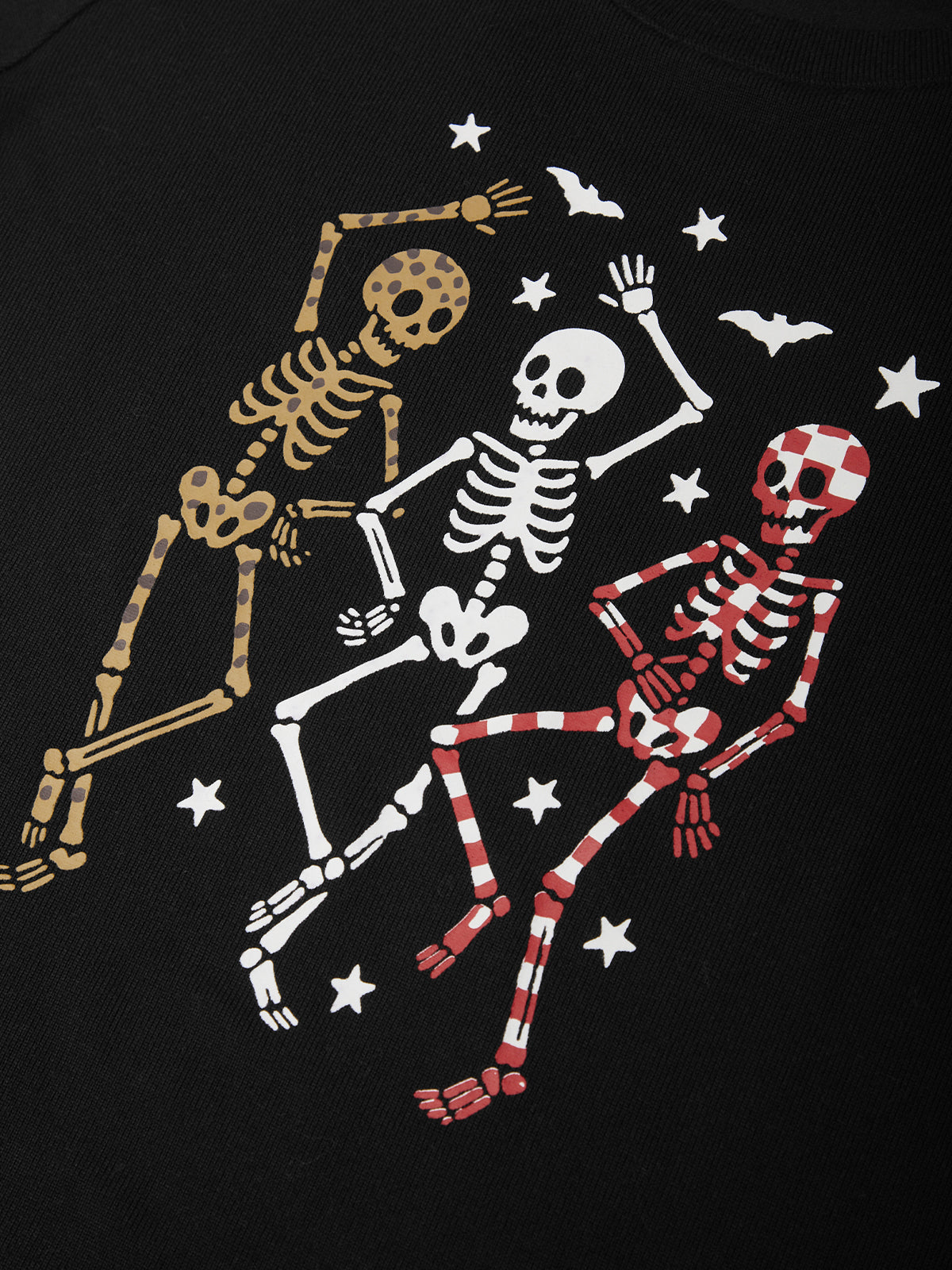 A close-up of a black garment featuring a graphic print of three dancing skeletons in gold, white, and red-and-white checkered patterns, surrounded by white stars and small bats.