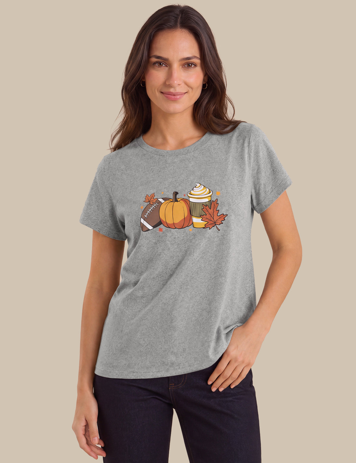 A woman modeling a heather gray short-sleeve t-shirt with a colorful autumn-themed graphic on the chest, featuring a pumpkin, football, hot dog, and fall leaves, set against a solid beige background.