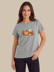 A woman modeling a heather gray short-sleeve t-shirt with a colorful autumn-themed graphic on the chest, featuring a pumpkin, football, hot dog, and fall leaves, set against a solid beige background.