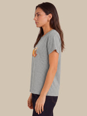 A woman shown in profile wearing a light heather gray short-sleeve t-shirt with a small orange and yellow graphic on the left chest, paired with dark pants, against a solid beige background.