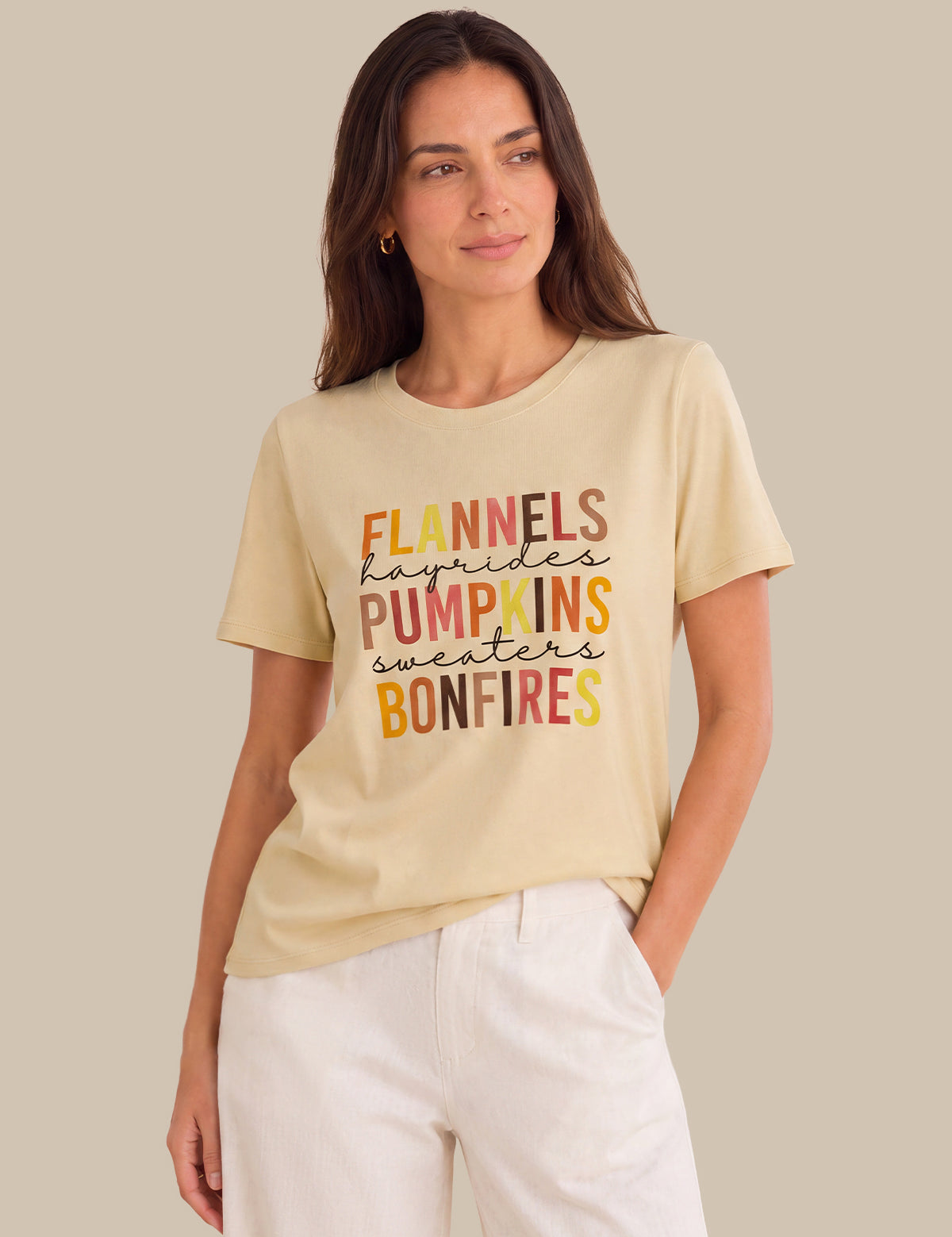 A woman wearing a beige graphic t-shirt with autumn-themed text and white pants, standing against a solid beige background.