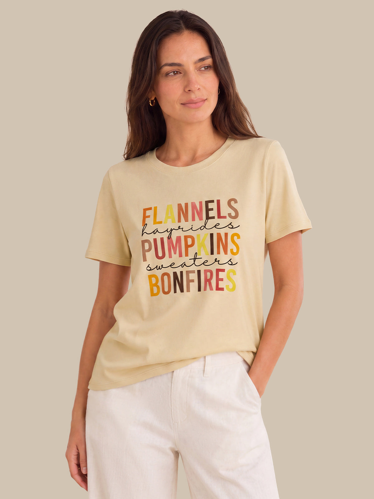 A woman wearing a beige graphic t-shirt with autumn-themed text and white pants, standing against a solid beige background.