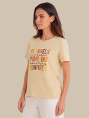 A woman wearing a pale beige graphic t-shirt with colorful text that reads 'FLANNELS humbles PUMPSINS sweaters BONFIRES', paired with white pants, set against a solid beige background.