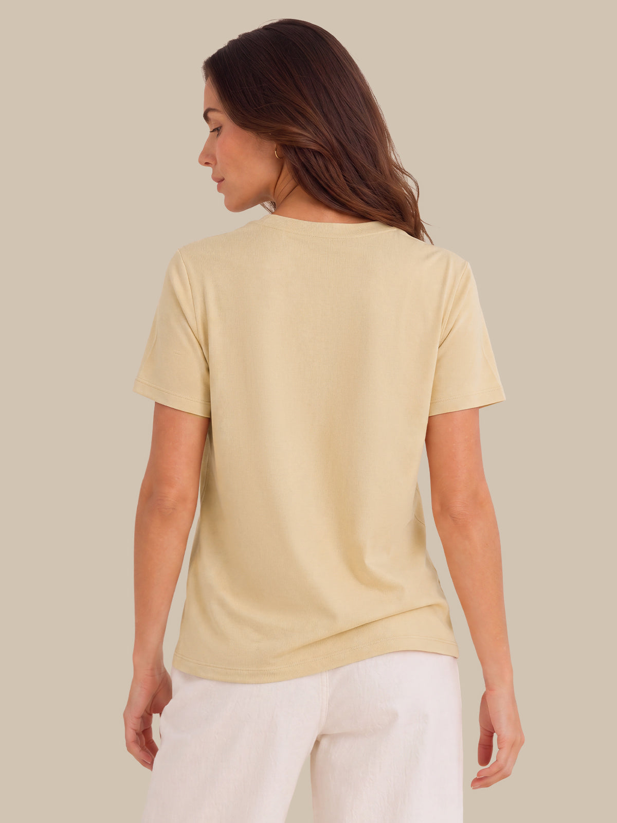 A woman shown from the back wearing a pale cream-colored short-sleeve t-shirt and white pants, set against a solid light beige background.