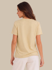A woman shown from the back wearing a pale cream-colored short-sleeve t-shirt and white pants, set against a solid light beige background.