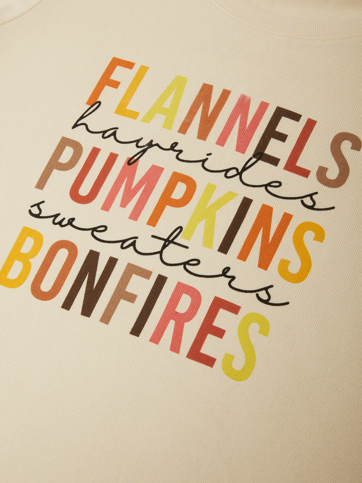 Close-up of a cream-colored sweatshirt featuring a colorful graphic text print that reads 'FLANNELS hayrides PUMPKINS sweaters BONFIRES' in orange, yellow, brown, and red lettering.