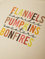 Close-up of a cream-colored sweatshirt featuring a colorful graphic text print that reads 'FLANNELS hayrides PUMPKINS sweaters BONFIRES' in orange, yellow, brown, and red lettering.