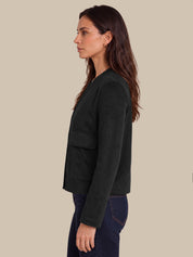 A woman shown in side profile wearing a black cropped jacket with flap pockets and dark blue jeans, set against a solid beige background.