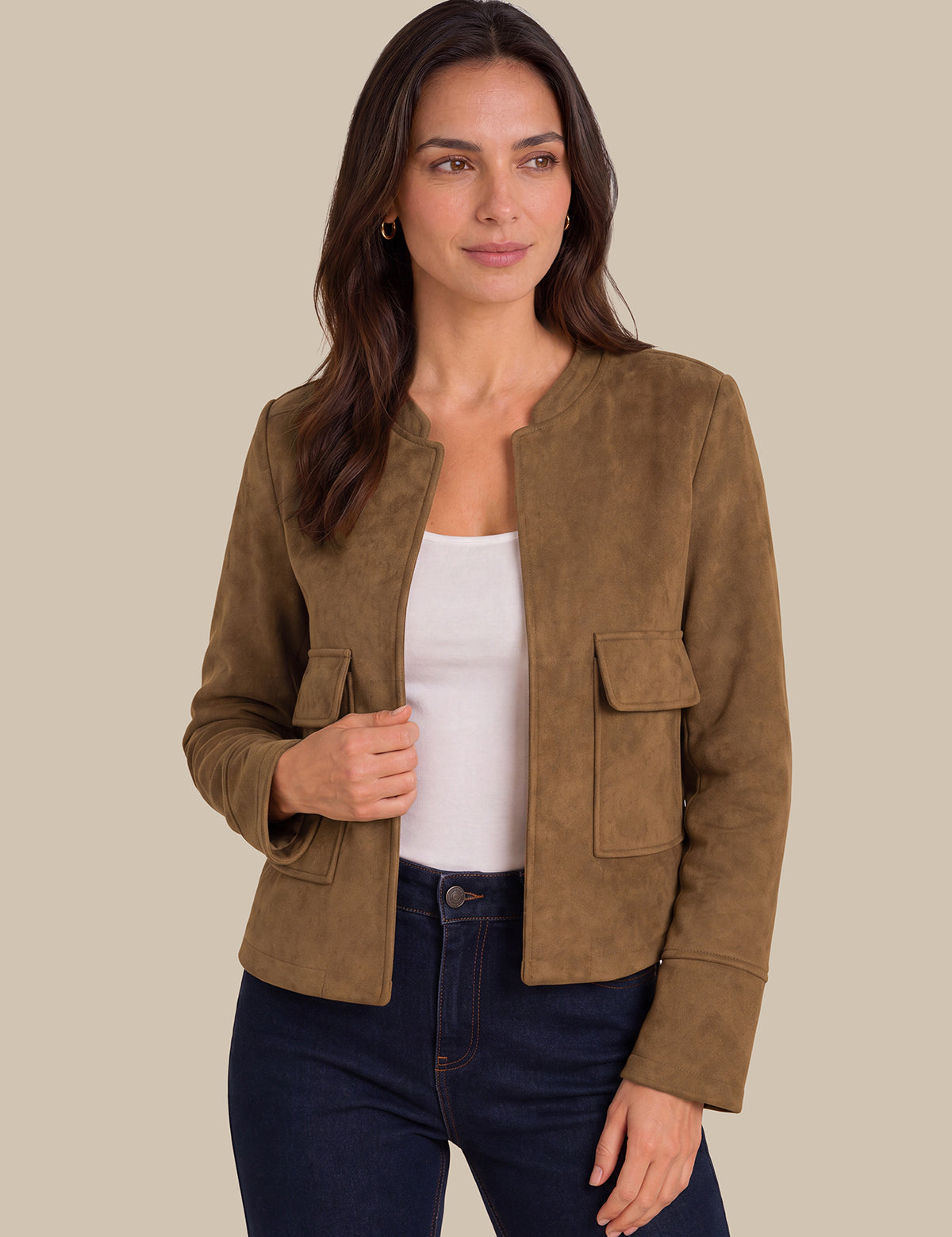 A woman modeling a cropped brown suede jacket over a white tank top and dark blue jeans, set against a solid beige background.