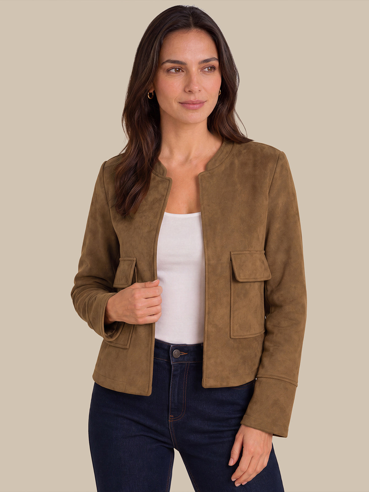 A woman modeling a cropped brown suede jacket over a white tank top and dark blue jeans, set against a solid beige background.