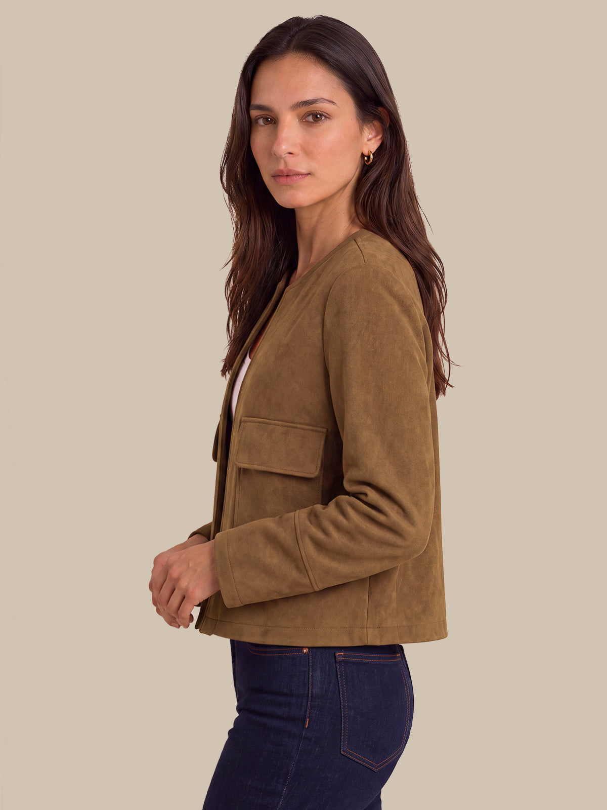 A woman wearing a cropped, long-sleeve suede jacket in a warm camel brown color, paired with dark blue jeans, shown from a side profile against a solid beige background.