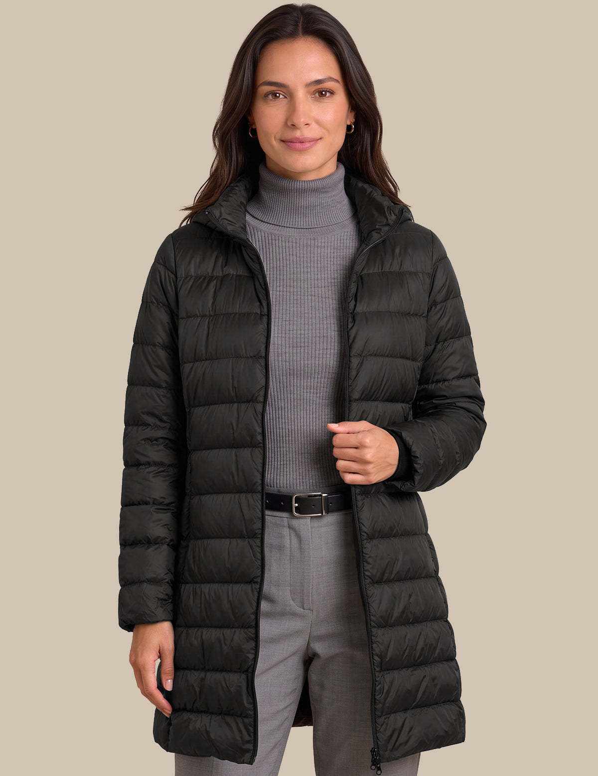 A woman modeling a black mid-length puffer coat over a gray ribbed turtleneck sweater and gray trousers, set against a solid beige background.