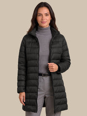 A woman modeling a black mid-length puffer coat over a gray ribbed turtleneck sweater and gray trousers, set against a solid beige background.