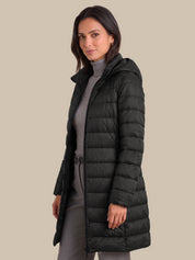 A woman modeling a long black hooded puffer coat over a gray turtleneck and gray skirt, photographed against a solid beige studio background.