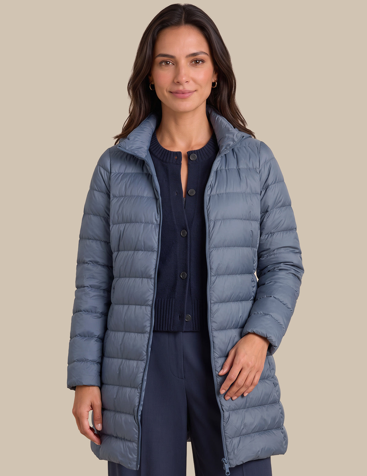 A woman modeling a long, slate blue-gray puffer jacket over a navy cardigan and navy pants, set against a solid beige background.