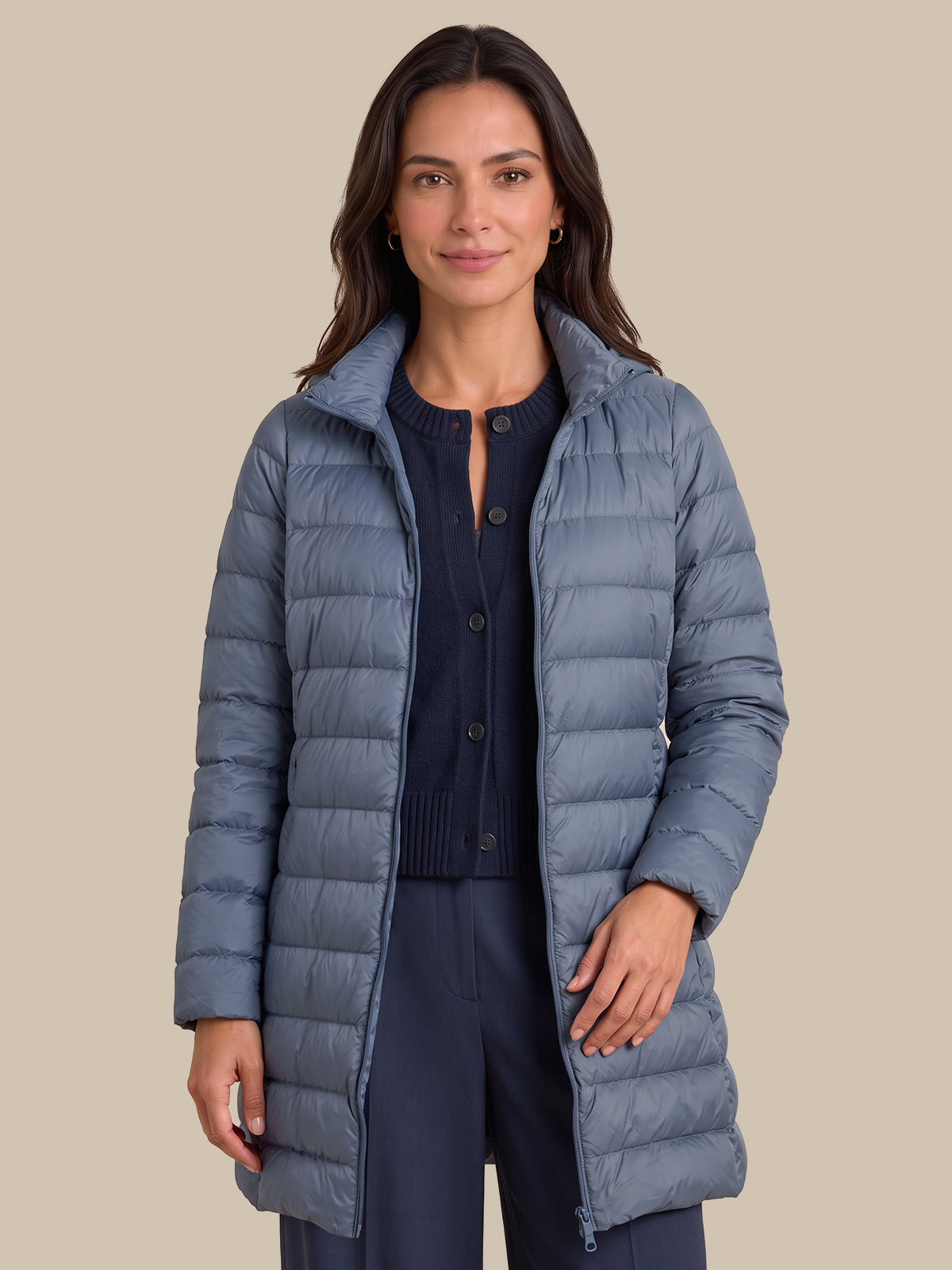 A woman modeling a long, slate blue-gray puffer jacket over a navy cardigan and navy pants, set against a solid beige background.