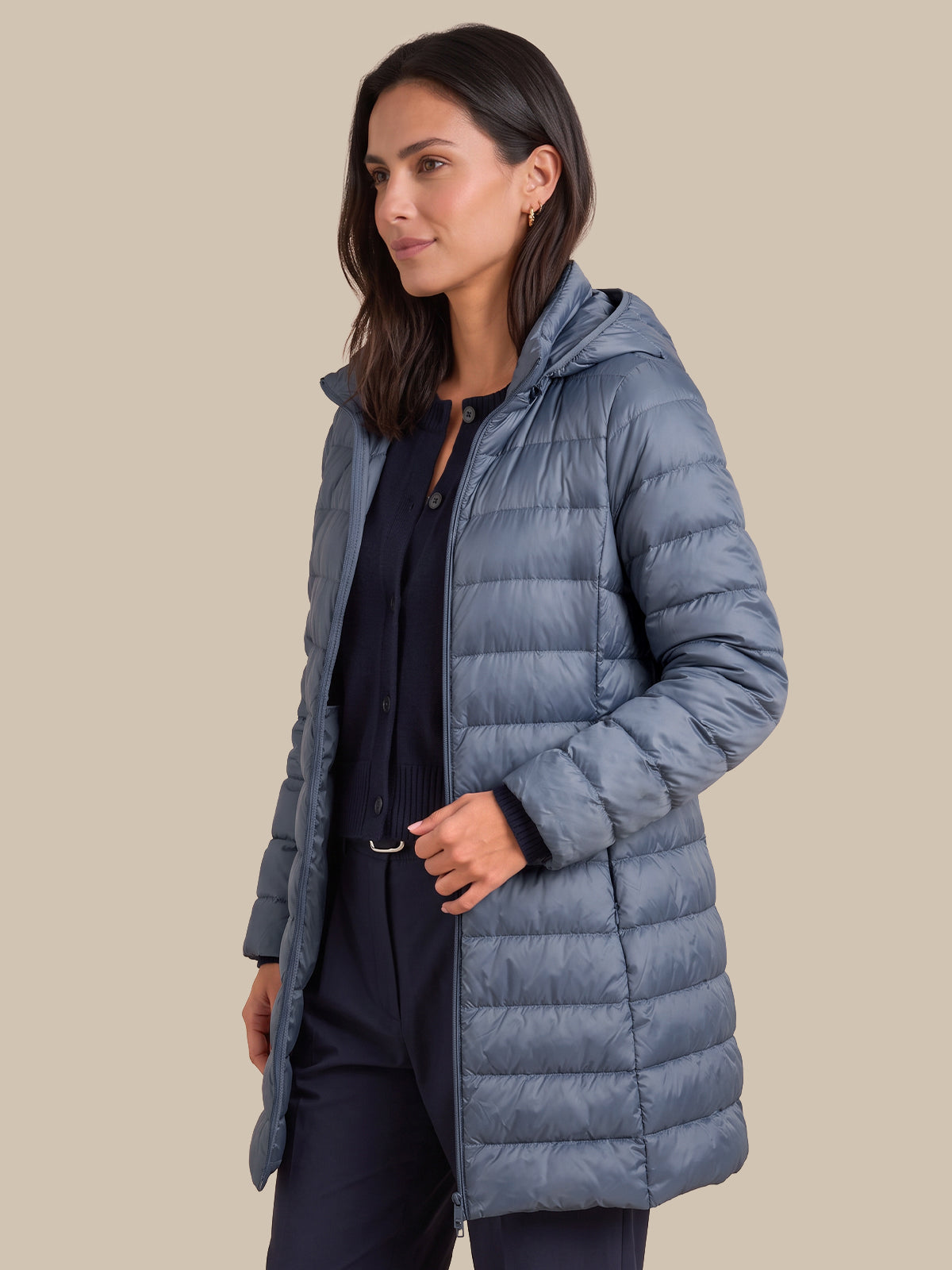 A woman wearing a mid-length, hooded puffer jacket in a dusty slate blue color, styled over a dark navy top and trousers, set against a solid beige background.