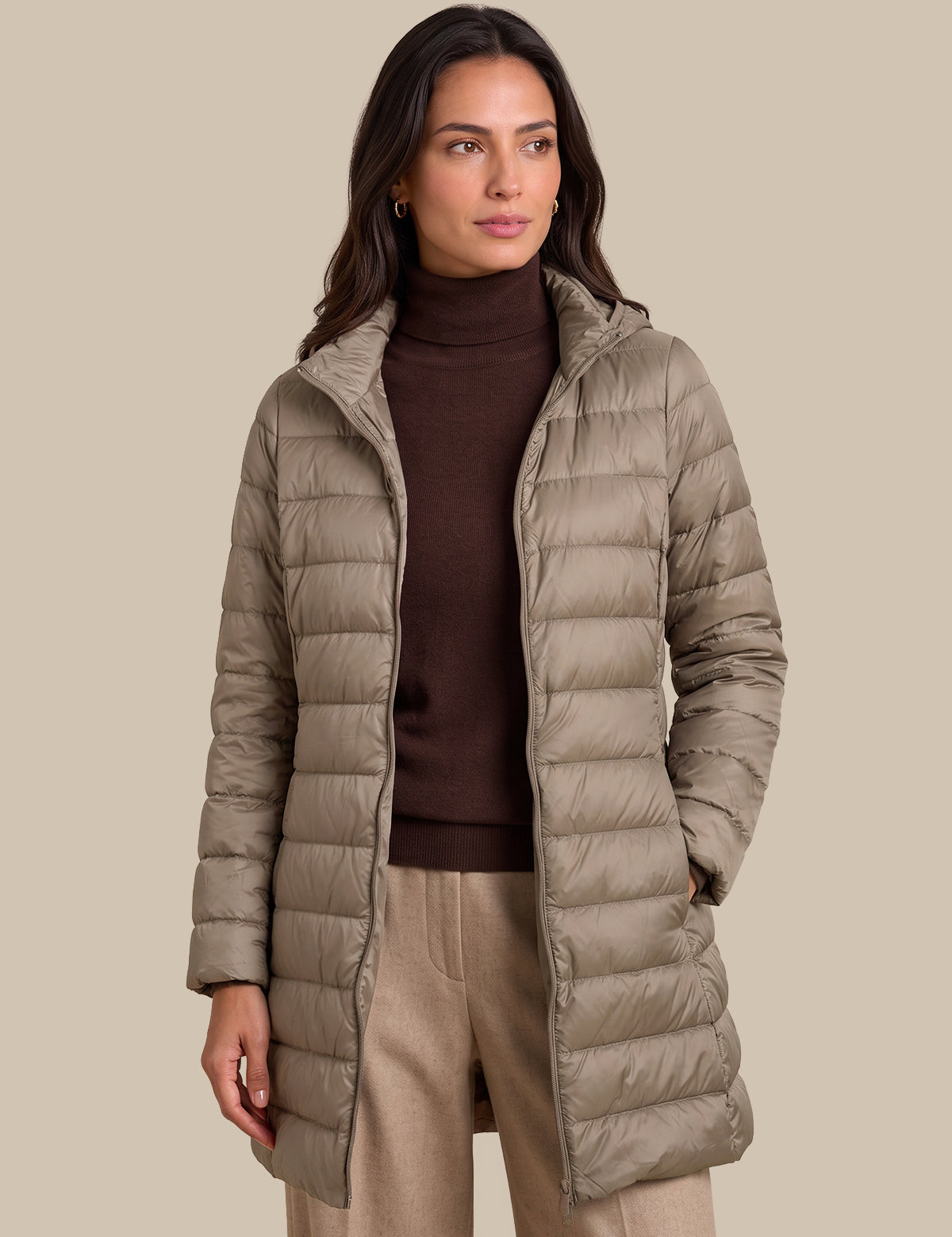 A woman wearing a mid-length taupe quilted puffer coat over a dark brown turtleneck sweater and beige trousers, standing against a solid beige background.