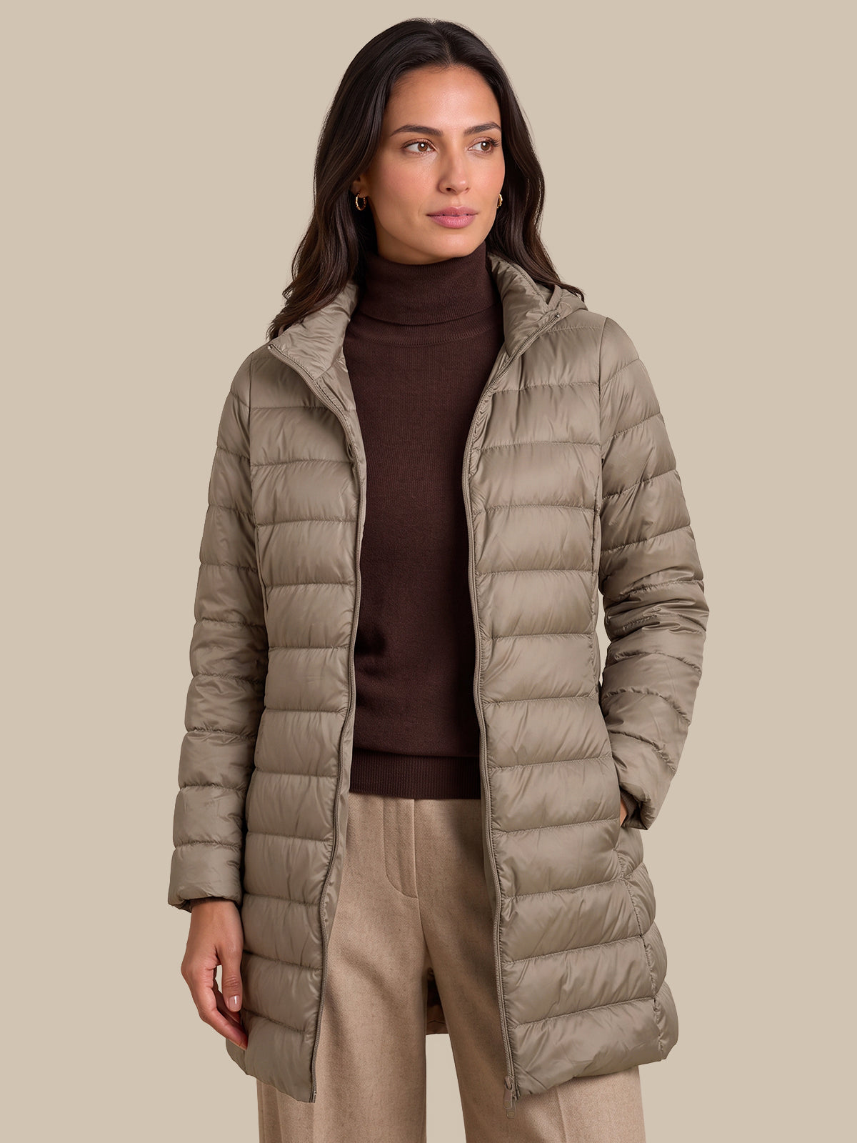 A woman wearing a mid-length taupe quilted puffer coat over a dark brown turtleneck sweater and beige trousers, standing against a solid beige background.