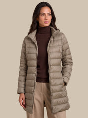 A woman wearing a mid-length taupe quilted puffer coat over a dark brown turtleneck sweater and beige trousers, standing against a solid beige background.