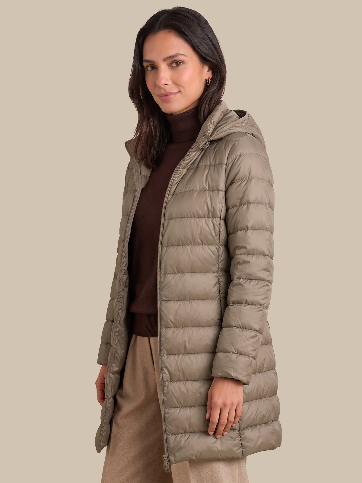 A woman modeling a long, light taupe-brown hooded puffer jacket over a dark brown turtleneck sweater and beige trousers, set against a solid beige background.