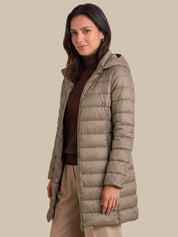 A woman modeling a long, light taupe-brown hooded puffer jacket over a dark brown turtleneck sweater and beige trousers, set against a solid beige background.