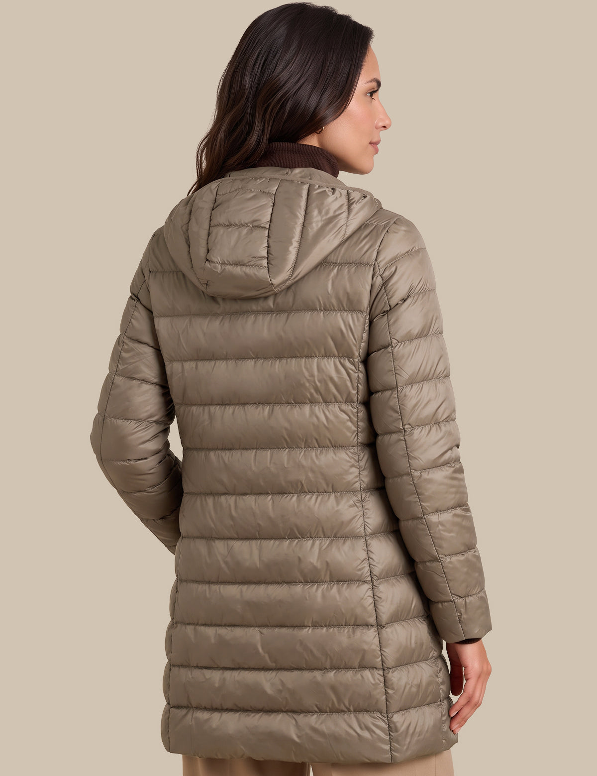 A woman shown from the back wearing a hooded puffer jacket in a taupe-beige color, photographed against a solid light beige studio background.
