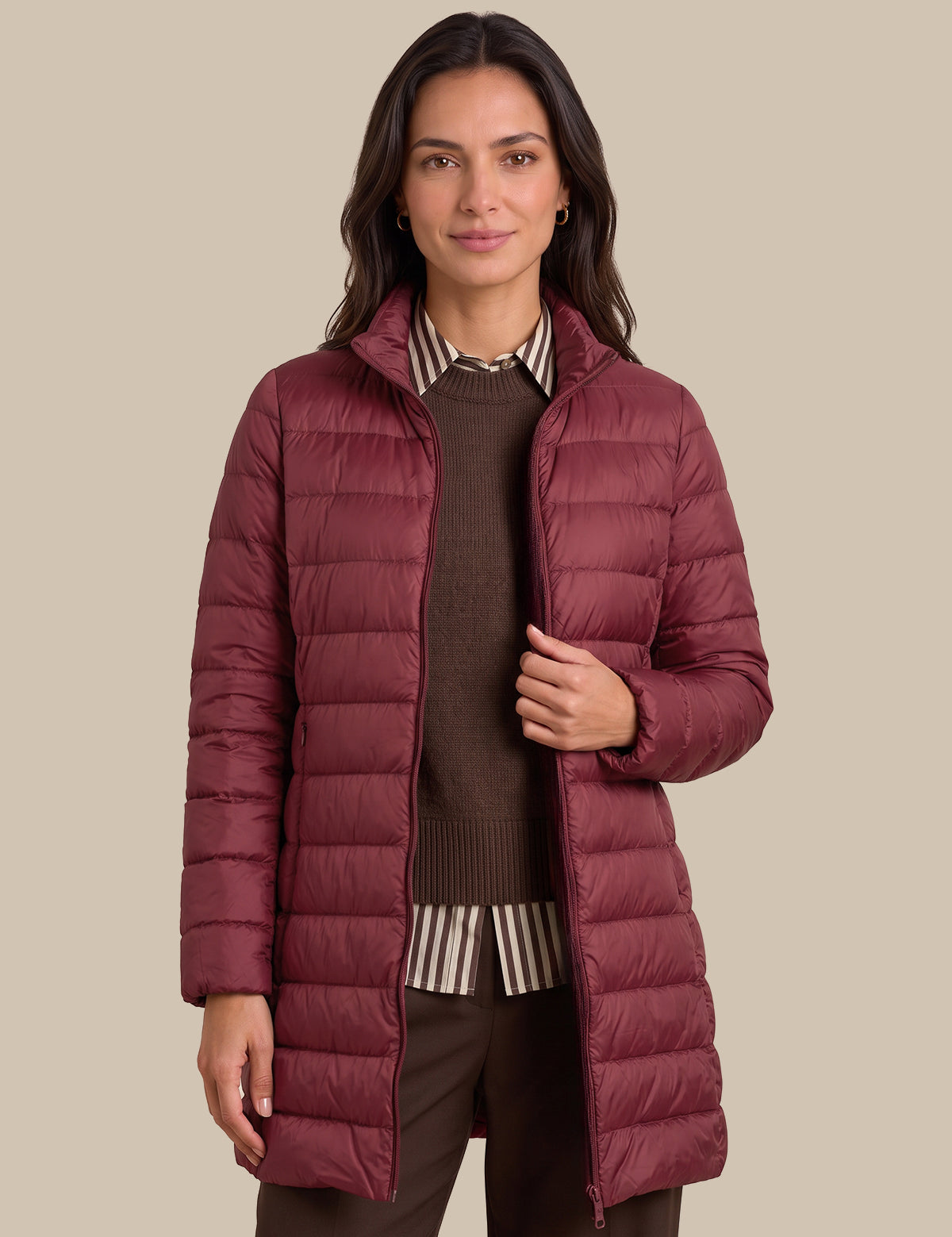 A woman modeling a long, quilted puffer jacket in a deep burgundy color, worn over a brown sweater and striped shirt, against a solid beige background.