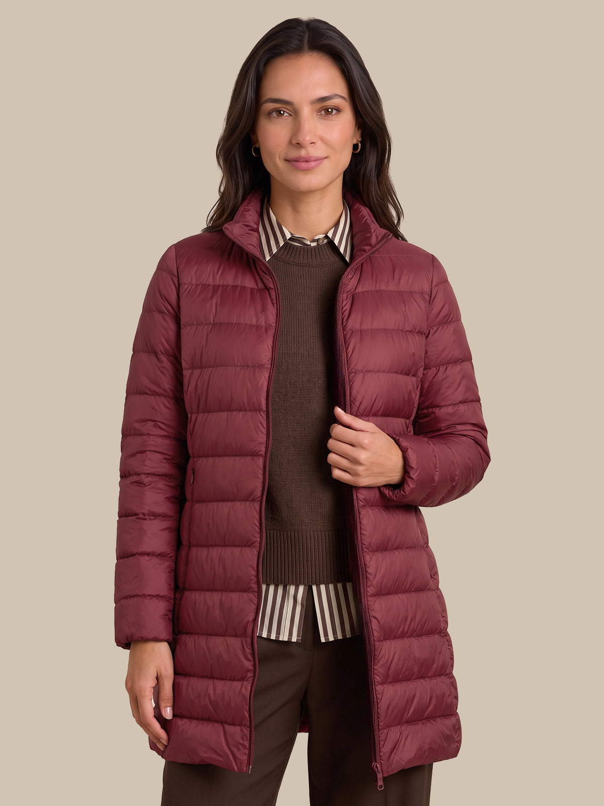 A woman modeling a long, quilted puffer jacket in a deep burgundy color, worn over a brown sweater and striped shirt, against a solid beige background.