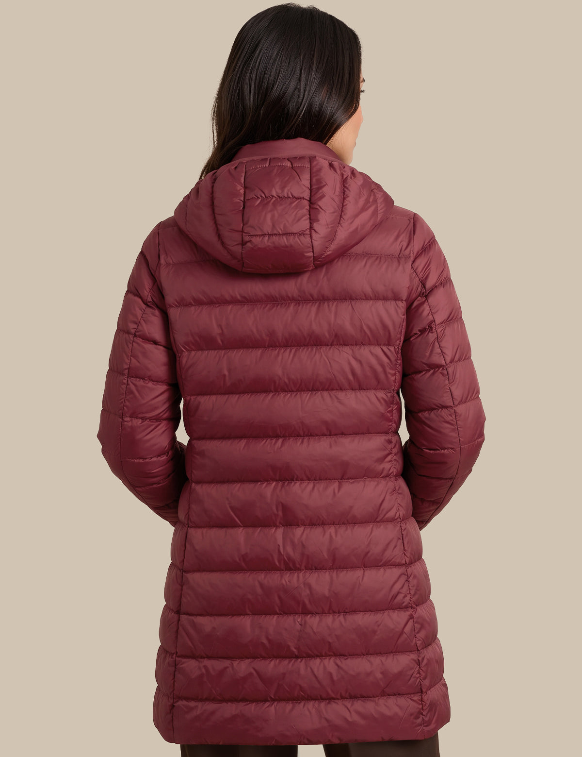 A woman viewed from the back wearing a long quilted puffer coat in a muted burgundy or burgundy-rose tone, against a plain warm beige studio background.
