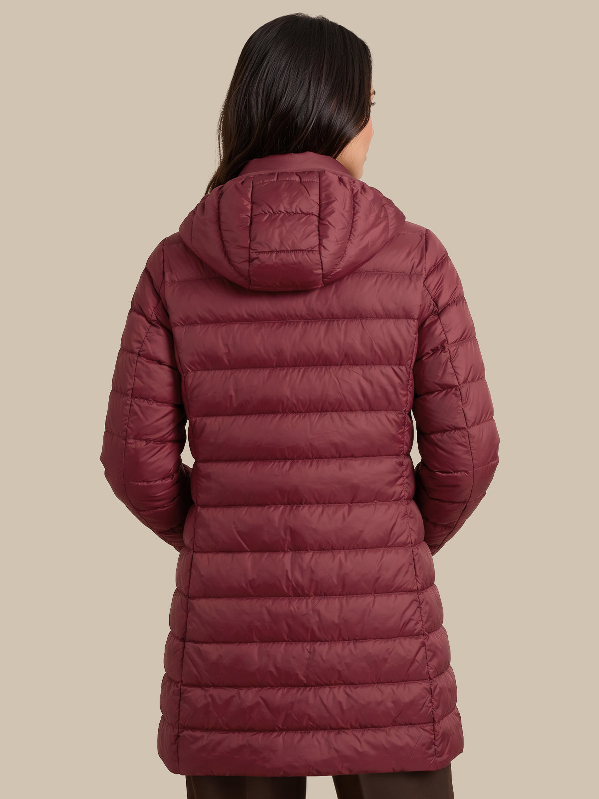 A woman viewed from the back wearing a long quilted puffer coat in a muted burgundy or burgundy-rose tone, against a plain warm beige studio background.