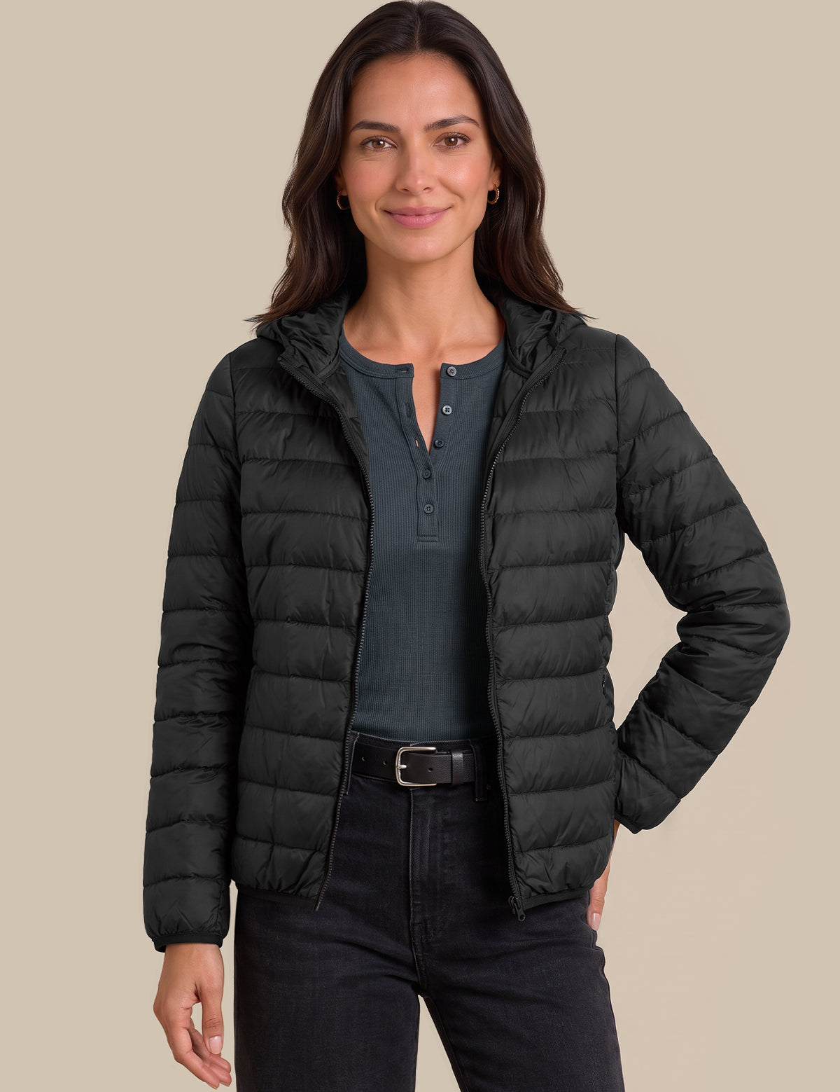 A woman modeling a black hooded puffer jacket over a dark gray henley shirt and black jeans, set against a solid beige background.