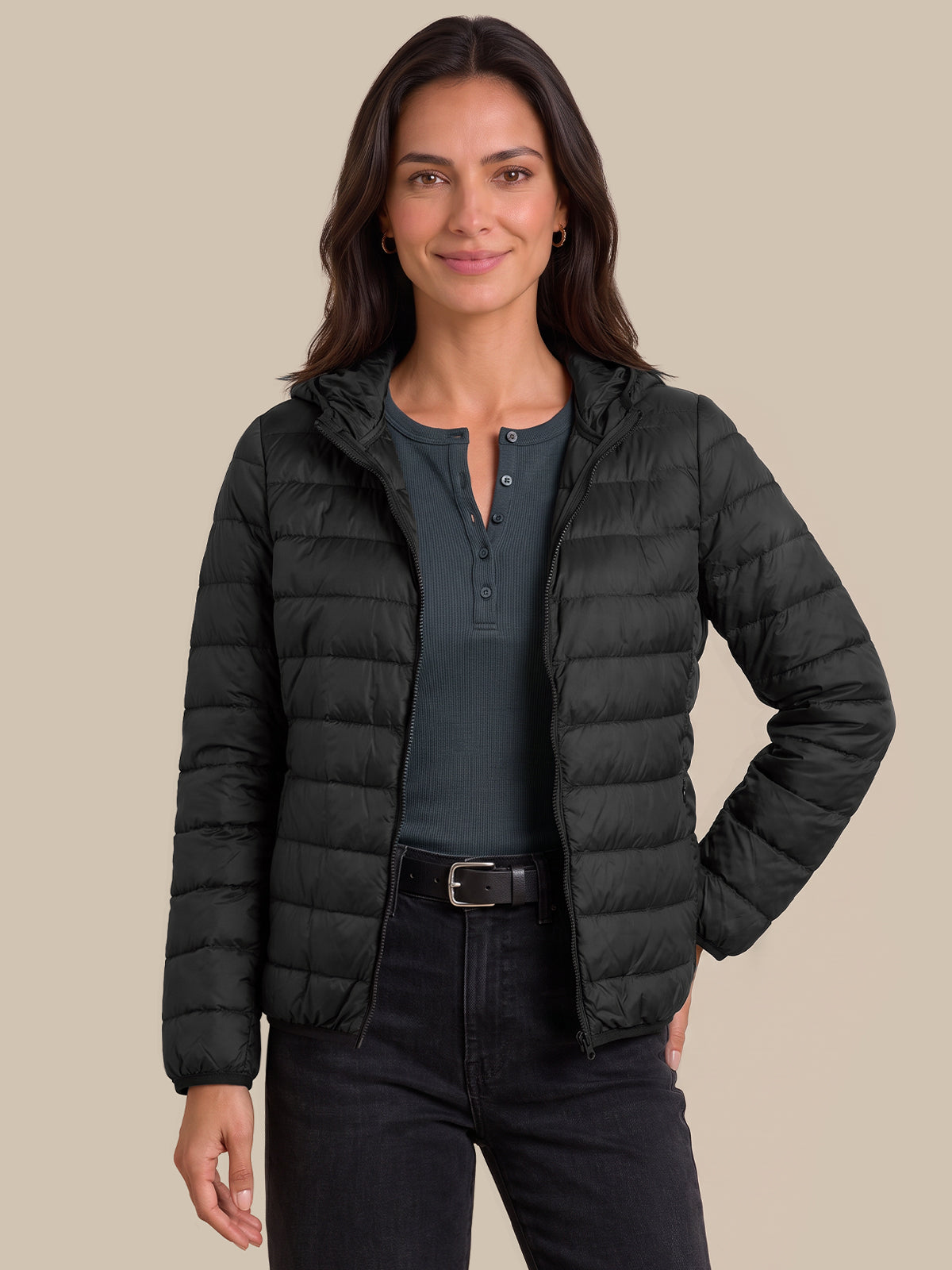 A woman modeling a black hooded puffer jacket over a dark gray henley shirt and black jeans, set against a solid beige background.