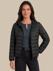 A woman modeling a black hooded puffer jacket over a dark gray henley shirt and black jeans, set against a solid beige background.
