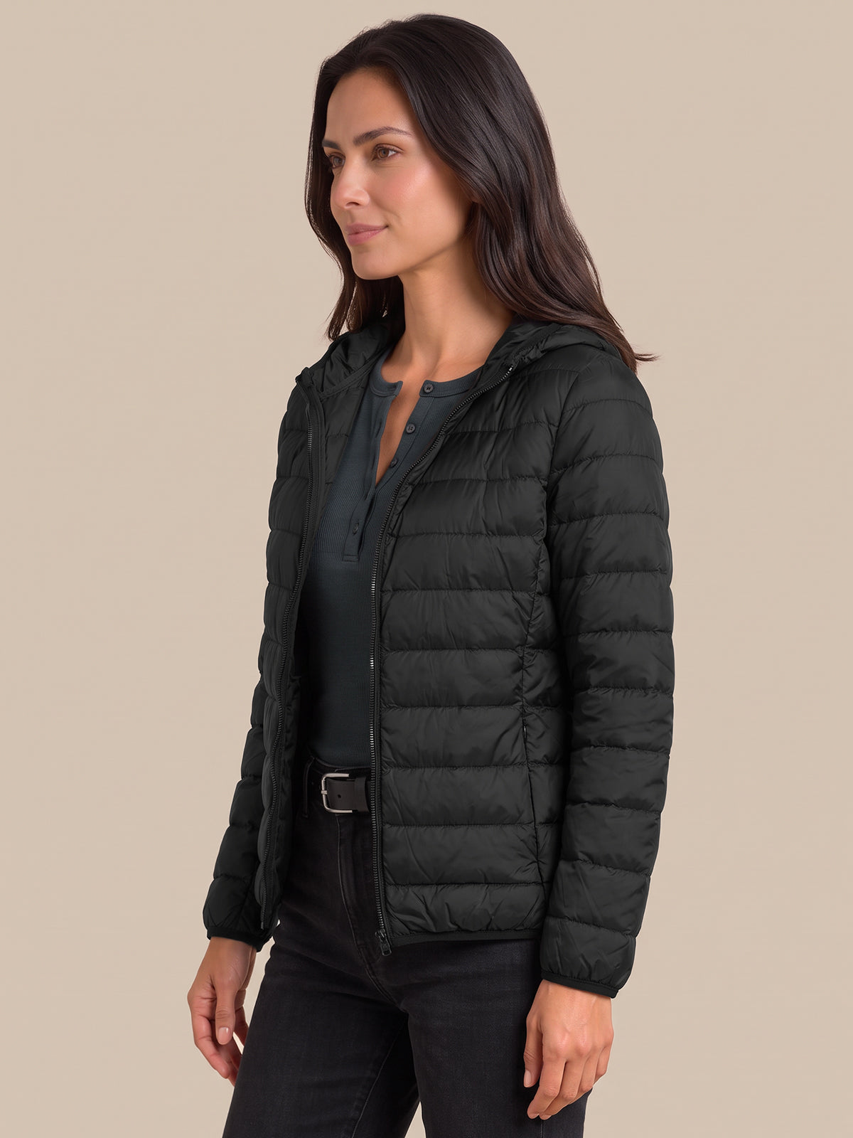 A woman wearing a black hooded puffer jacket over a dark top and black pants, photographed against a plain beige studio background.