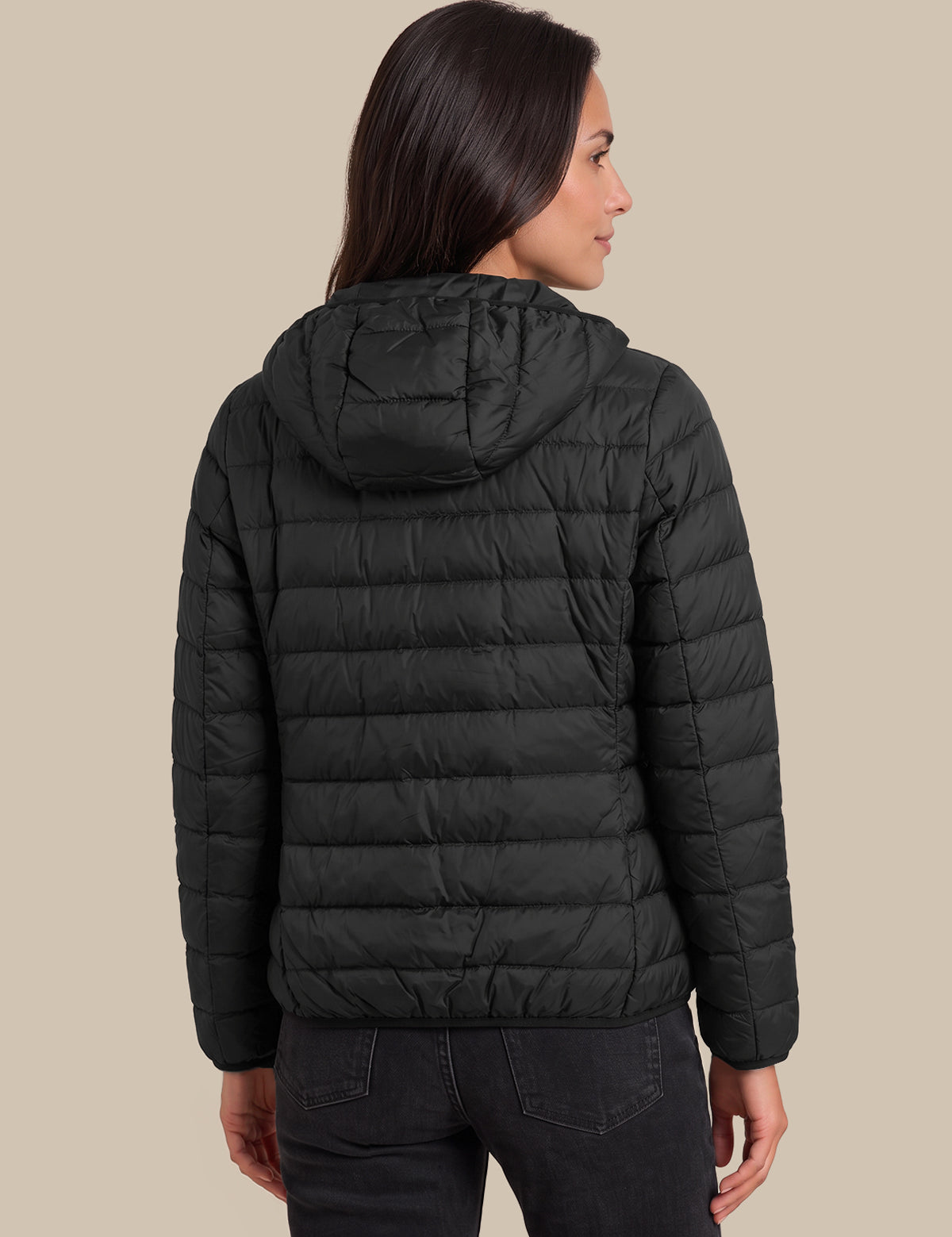 A woman shown from the back wearing a black hooded puffer jacket and dark jeans against a solid beige studio background.