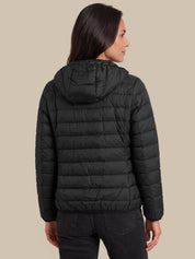 A woman shown from the back wearing a black hooded puffer jacket and dark jeans against a solid beige studio background.