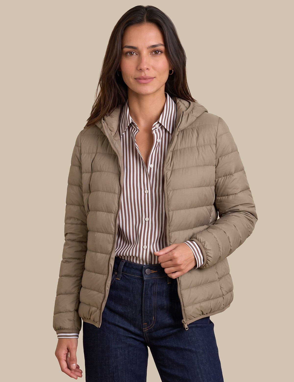 A woman modeling a light taupe hooded puffer jacket over a brown and white striped button-down shirt and dark blue jeans, set against a solid beige background.