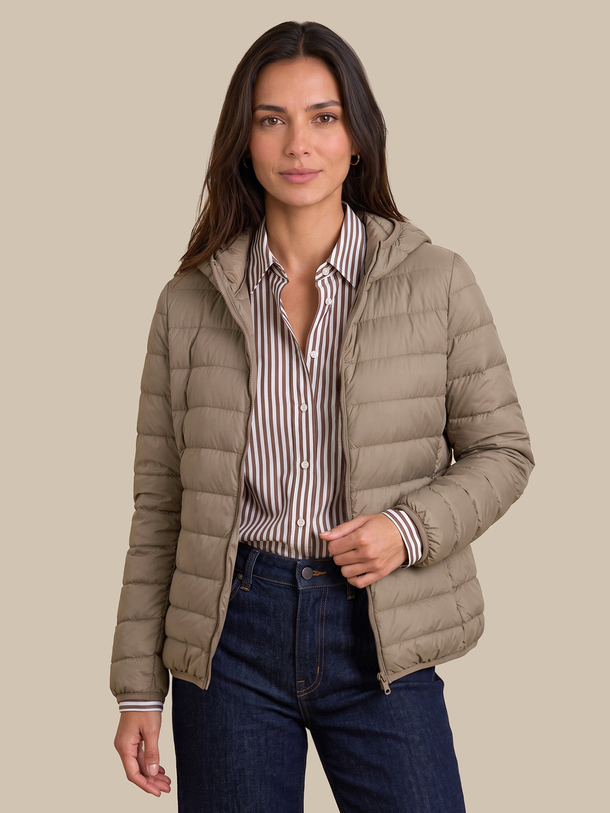 A woman modeling a light taupe hooded puffer jacket over a brown and white striped button-down shirt and dark blue jeans, set against a solid beige background.