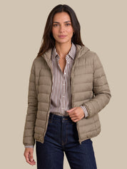 A woman modeling a light taupe hooded puffer jacket over a brown and white striped button-down shirt and dark blue jeans, set against a solid beige background.