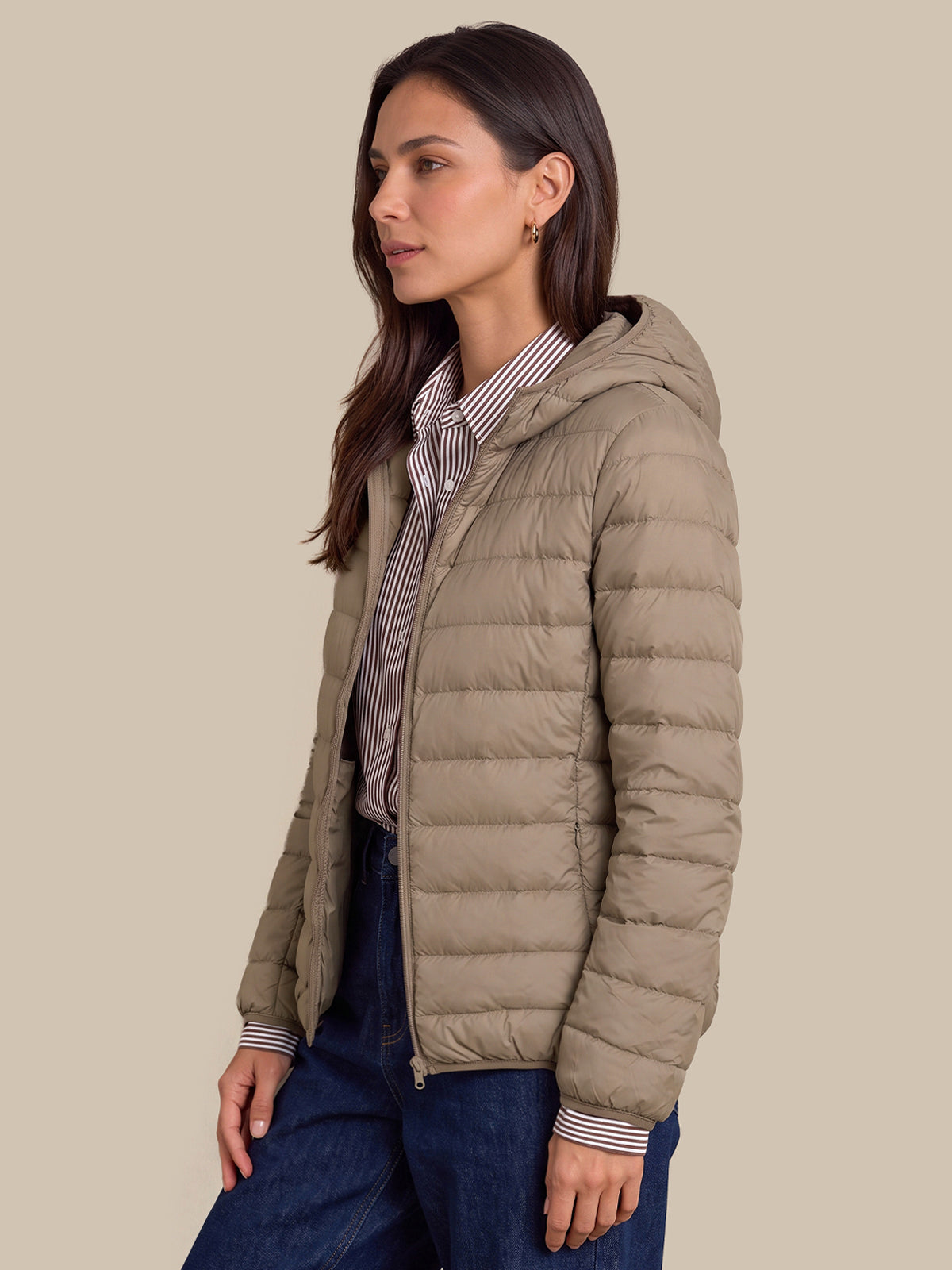 A woman wearing a light taupe hooded puffer jacket over a striped button-up shirt and dark blue jeans, photographed against a solid beige studio background.