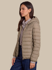 A woman wearing a light taupe hooded puffer jacket over a striped button-up shirt and dark blue jeans, photographed against a solid beige studio background.