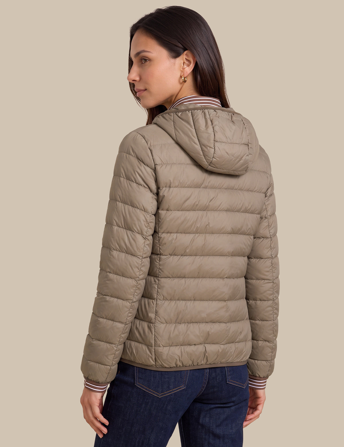 A woman shown from the back wearing a light taupe quilted puffer jacket with a hood, paired with dark blue jeans, against a solid beige background.
